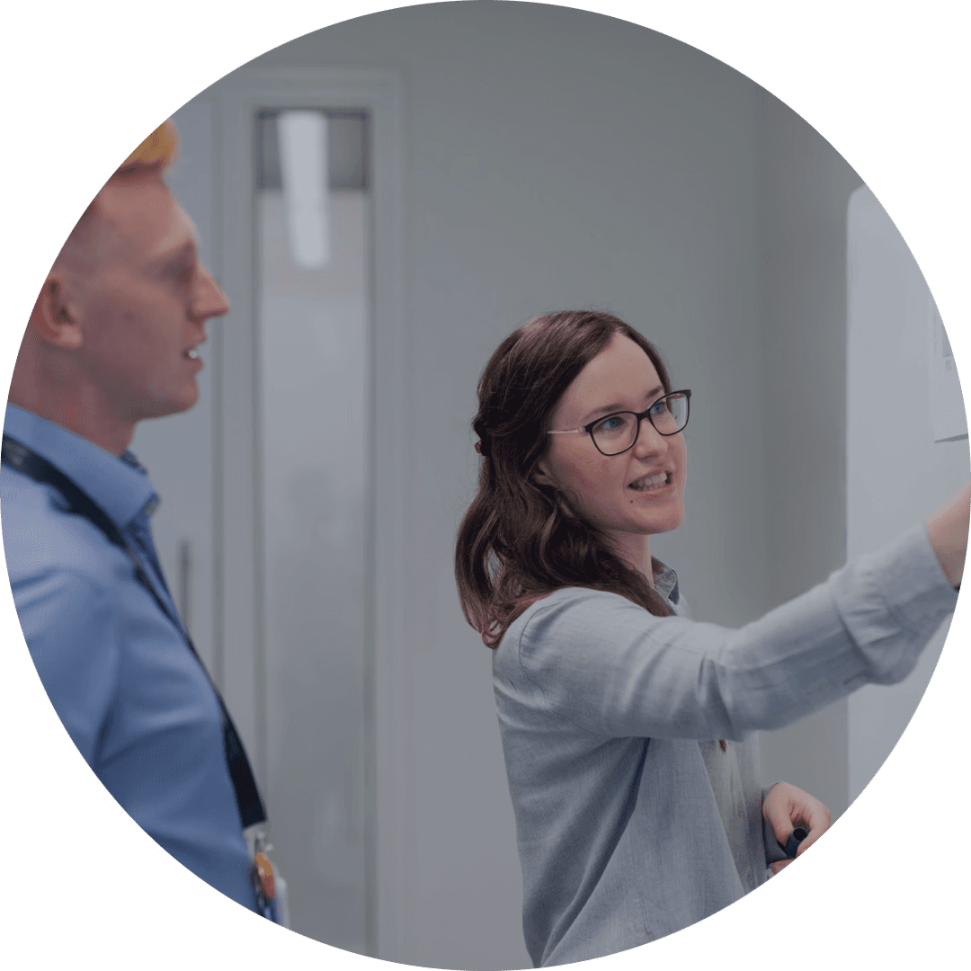 Business professionals collaborating in office setting - woman with dark brown hair and glasses pointing/writing on board, man with light red/blonde hair in blue shirt listening attentively